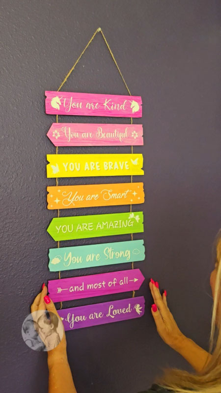 Rainbow nursery wall décor hanging against a purple wall. The sign unfolds into eight connected wooden panels with inspirational quotes such as “You are brave, you are smart.” Made of sturdy wood with pink and rainbow tones, shown hanging by a rope in a nursery.