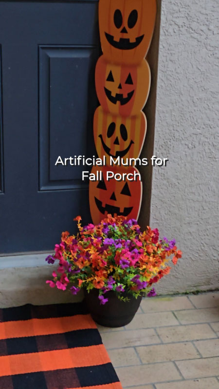 Close-up of artificial fall mums in orange, purple, and rose red colors, displayed in a planter by the porch.
