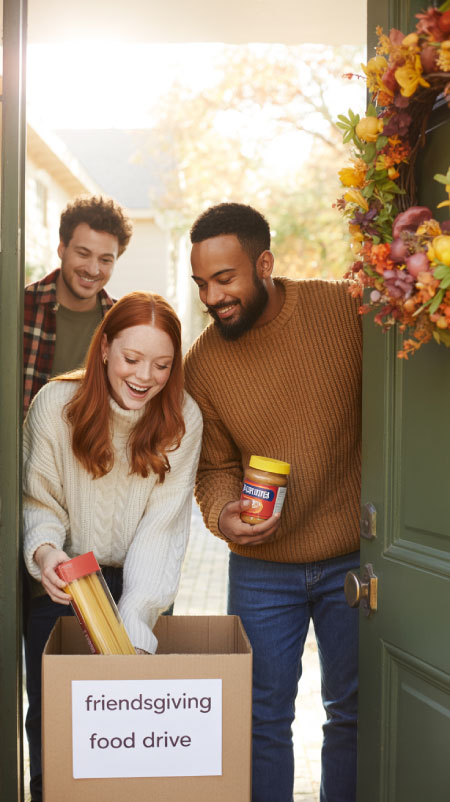 Friends placing canned goods and wrapped items into a “Friendsgiving Food Drive” donation box near a front door with a fall wreath. The moment captures generosity and gratitude as part of a Thanksgiving-with-friends tradition.