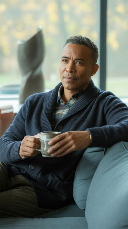 African-American man seated on a blue couch holding a cup of coffee, wearing a navy shawl-collar cardigan over a plaid button-down shirt; window behind him shows colorful autumn leaves — cozy Friendsgiving outfit idea for men.