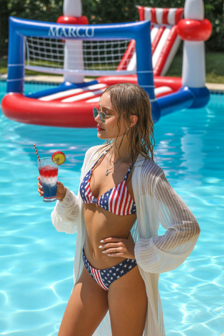 Woman wearing a patriotic swimsuit and white cover-up holding a red white and blue drink by a pool with USA party floats