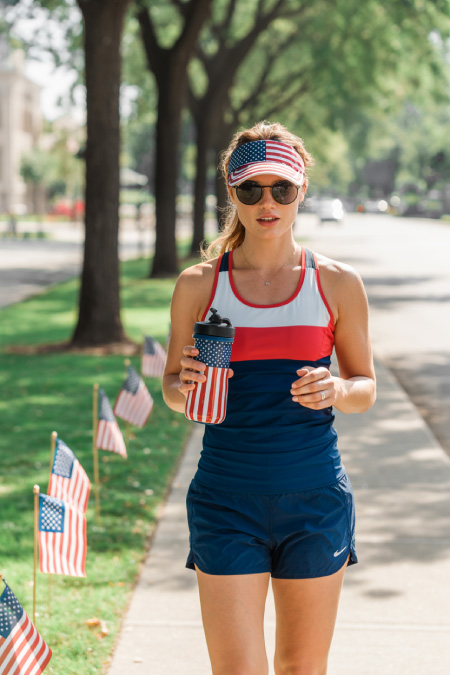 Woman wearing red white and blue athletic outfit walking outdoors with patriotic visor and tumbler during summer 4th of July celebration