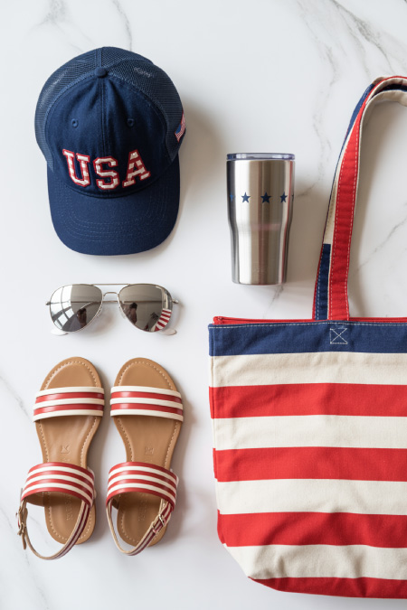 Patriotic outfit accessories including USA hat, striped tote bag, sandals, sunglasses, and tumbler styled flat lay for 4th of July outfit ideas
