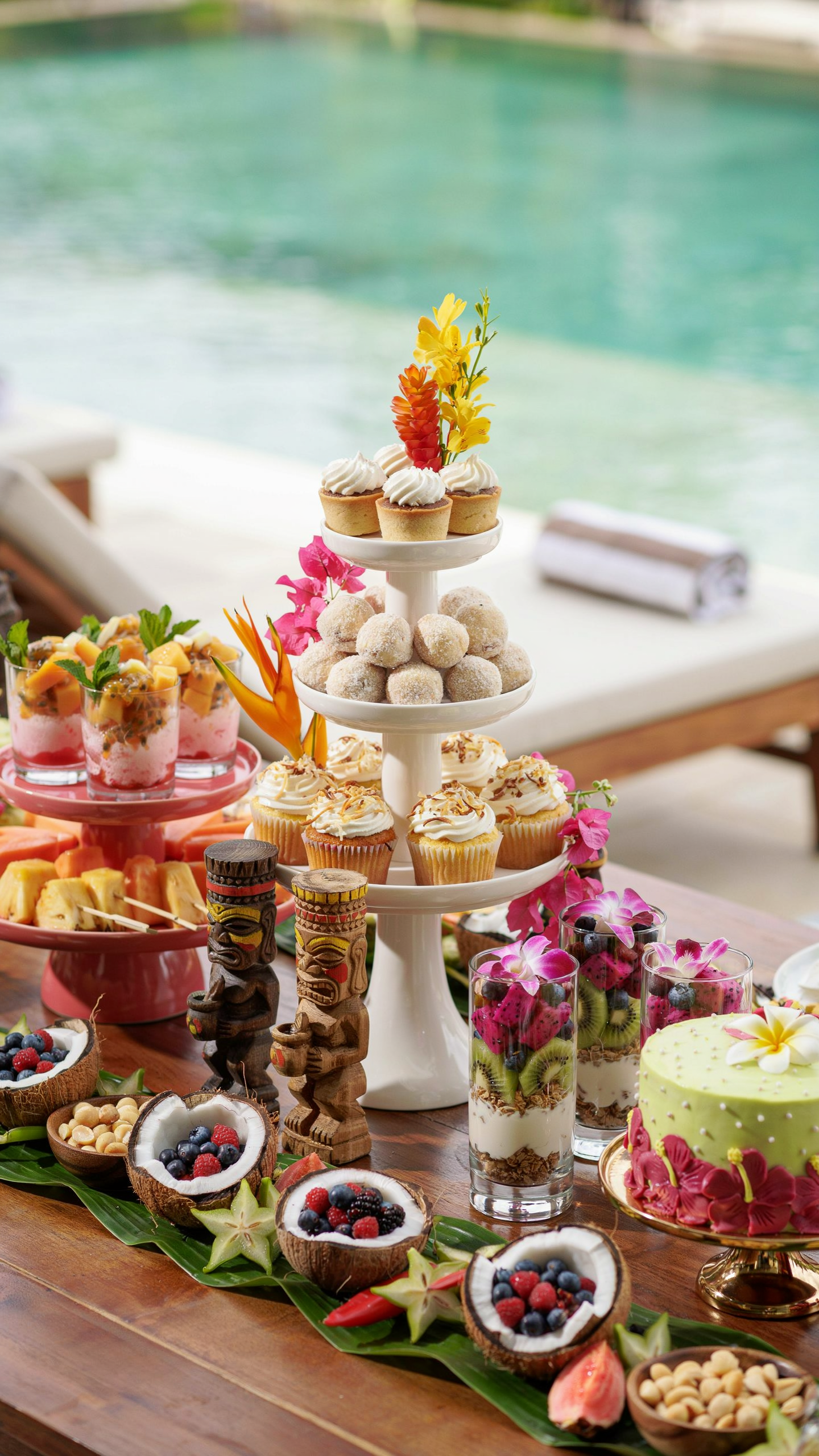 Tropical dessert station featuring cupcakes, mini desserts, tropical fruit parfaits, coconut bowls, decorated cake, tropical flowers, and tiki accents displayed beside a swimming pool.